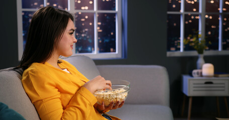 Happy smiling young woman sitting on couch at home watching funny tv programme comedy film, eating popcorn and laughing. Side view close up.