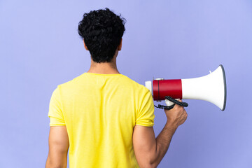 Young Venezuelan man isolated on purple background holding a megaphone and in back position