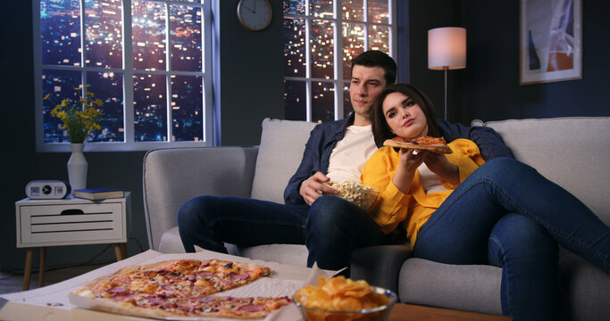 Caucasian Young Family Couple Watching Tv At Home, Sitting On Couch Snacking Popcorn And Eating Pizza. Loving Man And Woman Resting On Sofa Enjoying Movie Night.