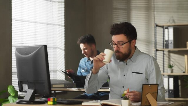 Young man in glasses and casual outfit eating sandwich while working in office