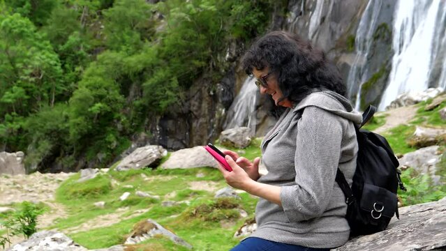 Woman sitting on the rock by waterfall looking at recent pictures on her mobile phone laughing and commenting excitedly.