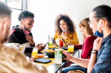 Happy multiracial friends lowering their masks and smile while using mobile phones sitting at restaurant - Young millennials influencers working at bar - Youth lifestyle and new normal concept