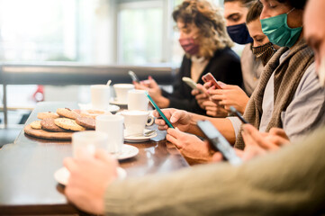 Obraz na płótnie Canvas Young influencer people working with smartphone sitting in a coffee shop while wearing protective face mask for coronavirus prevention - New normal lifestyle concept about teamwork, tech, startup
