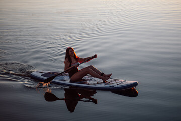 Blissful middle aged woman sitting on sup board rowing with oar on lake with clear blue water...