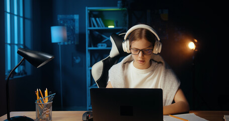Young teenage caucasian girl using laptop and headphones to study online, sitting in dark cozy room at home. Clever teenager learning language with internet tutor via zoom educational platform.
