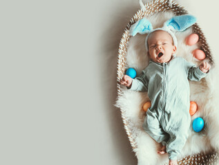Fototapeta premium Cute baby Easter bunny. Little baby boy with bunny ears and Easter eggs in wicker basket in white fur. Symbol of Easter holiday, birth, spring, religion.