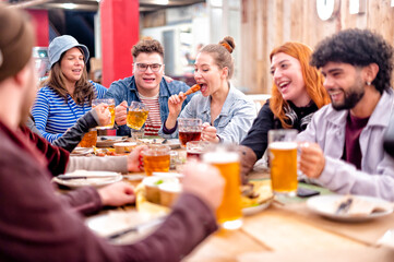 Happy girl sitting at the wooden table drinks beer and eats chicken leg together with friends at restaurant bar - Group of cheerful young friends meeting for drink and eat food making a toast at pub