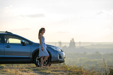 Happy young woman driver in blue dress leaning on her car enjoying warm summer day. Travelling and vacation concept