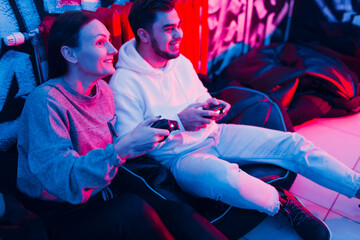 Young man and woman couple plays on game console with gamepad sitting on chair.