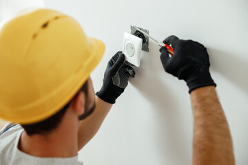 Professional electrician in uniform using screwdriver while installing electrical socket outlet...