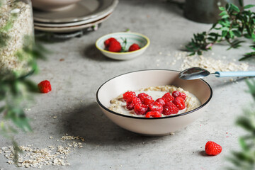 Bowl of porridge with oats and raspberries at grey concrete kitchen table with branches and plates. Healthy vegan breakfast with seasonal summer berry. Front view.
