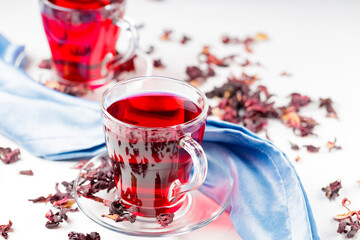 Hibiscus tea in glass cup on a blue napkin. Cup of red hibiscus tea and dry hibiscus petals on a white background. Natural dietary herbal tea