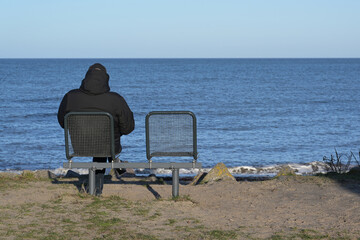 Fototapeta premium Single man in a dark warm jacket from behind sitting next to an empty chair on the sea shore looking over the water to the horizon, recreation or mourning for loss of a loved person, copy space