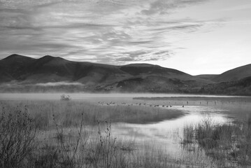 Black and white Absolutely stunning vibrant Autumn sunrise landscape image looking from Manesty Park in Lake Distict towards sunlit Skiddaw Range with mit rolling across Derwentwater surface