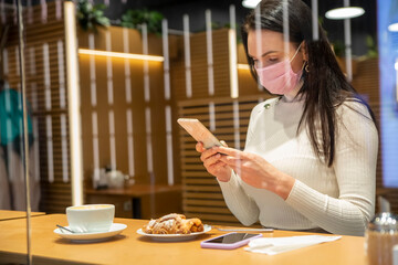 view through shop window of the cafe as a beautiful woman in a medical mask Sits and reads news in phone