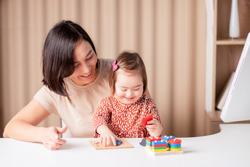 happy child girl with down syndrome with mom with educational toys, the kid at the table is studying