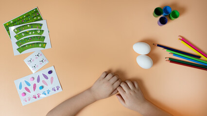 Children's hands make an Easter decoration on the egg. Pencils and stickers on a yellow background. Easter concept. Copy space.