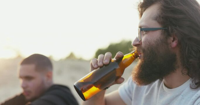 Face of a man with long hair and thick beard wearing glasses is visible in profile boy is drinking beer from glass bottle and talking with friend sitting outside in background rays of setting sun glow