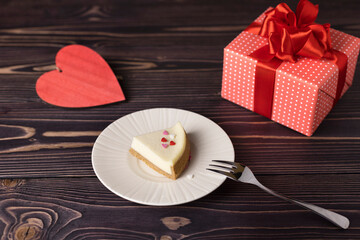 a classic piece of cheesecake on a white plate with a red heart and a gift on a wooden table. Selective focus.