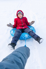 Fototapeta na wymiar Smiling boy lying on blue tubing in winter clothes on white snow and hand pulling rope and rolling him. Inflatable sleigh rides.