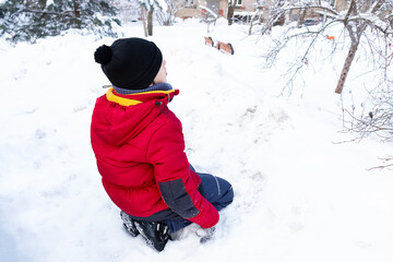 Fototapeta na wymiar Boy sitting in snow in winter clothes on snowbound street, view from back