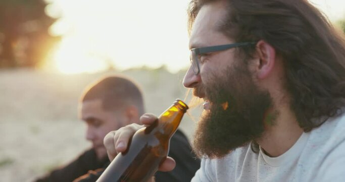 Face of a man with long hair and thick beard wearing glasses is visible in profile boy is drinking beer from glass bottle and talking with friend sitting outside in background rays of setting sun glow