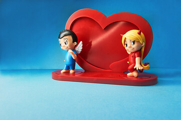 Toy model in the form of a boy angel and a girl devil next to the heart on a delicate blue background