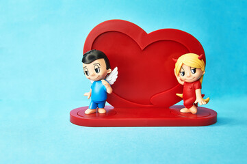 Toy model in the form of a boy angel and a girl devil next to the heart on a delicate blue background