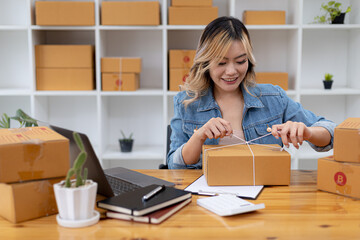 An Asian woman tying a parcel to a customer's box, she owns an online store, she packs and ships through a private transport company. Online selling and online shopping concepts.