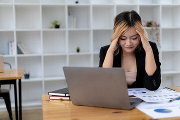 A businesswoman has a headache after working in front of her laptop for a long time and looking at a lot of paperwork causing stress, she is resting and working hard. Office syndrome concept.