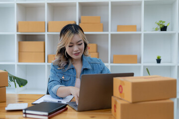 A beautiful Asian business owner opens an online store, she is checking orders from customers via laptop, sending goods through a courier company, concept of a woman opening an online business.