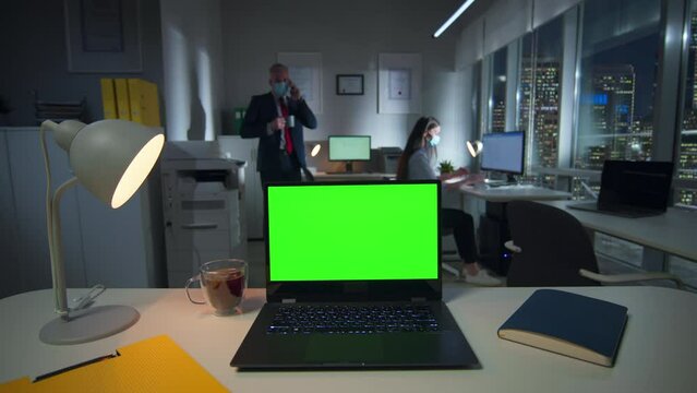 Laptop with green screen on desk and employees in safety mask working on background