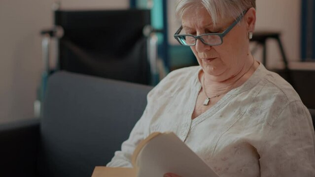 Retired woman reading literature book for knowledge in living room. Concentrated person enjoying novel story with fiction, resting on couch. Elderly adult with recreation hobby. Close up
