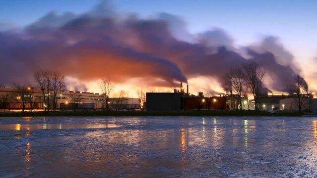 Emissions from industry fill the dawn sky with dark, unnatural clouds, reflected in ice covered river. 