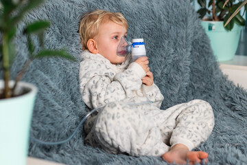 Cute baby boy makes inhalation with a nebulizer equipment. Sick child holding inhalator in hand and breathes through an inhaler at home