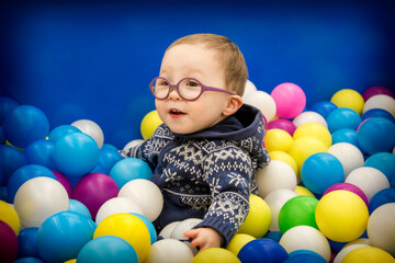 A little boy in a blue sweater with glasses plays with multi-colored plastic balls in the playroom.
