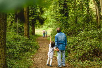 Girl and grandfather walk together on a path through green trees - obrazy, fototapety, plakaty