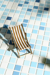 Concept of summer vacation. Miniature beach chair on swimming pool tile, sunlit light. Vertical orientation