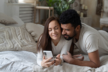 Smiling attractive couple using smartphone lying on bed together, happy guy and girlfriend checking social media news in morning, young woman showing man new mobile app