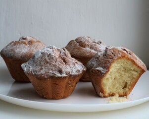 three whole muffins and one bitten one on a white plate, a set of homemade cakes close-up on a light background, a beautiful sweet pastry in a composition against a texture wall