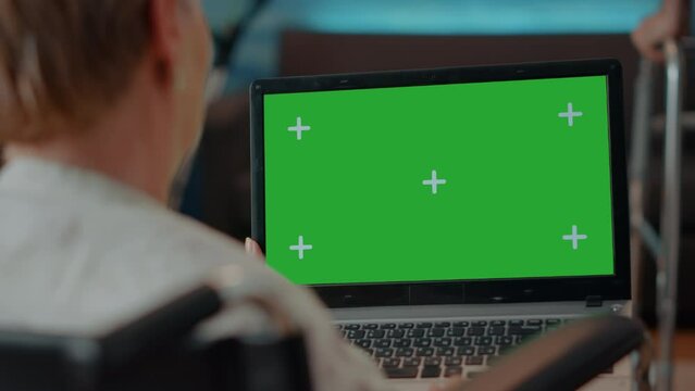 Elder person with disability using green screen on laptop, looking at isolated mock up template and blank copy space. Woman sitting in wheelchair with chroma key background on computer.