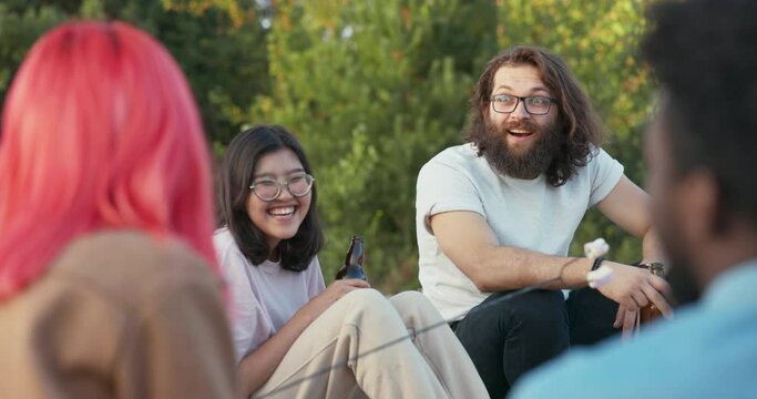 A man with long hair and beard wearing glasses sits next to a girl of another nationality, a beautiful woman of Asian beauty, they talk with friends exchange opinions, drink beer together