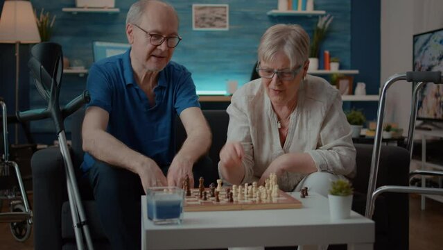 Senior couple playing chess board game with pieces, next to crutches and walking frame. People enjoying intellectual match with turn, king and pawn, thinking about moves to do checkmate.