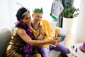 Happy black woman and her Caucasian friend wear Mardi Gras costumes and take selfie while getting...