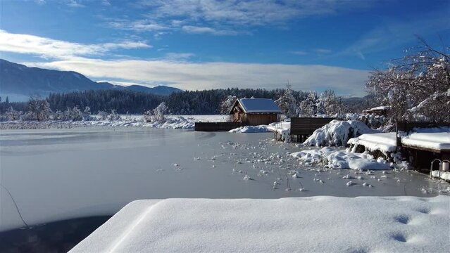 A panoramic view on the Faaker lake in Austrian Alps. The lake is surrounded by high mountains. There is a snow covered pier going into the frozen lake.