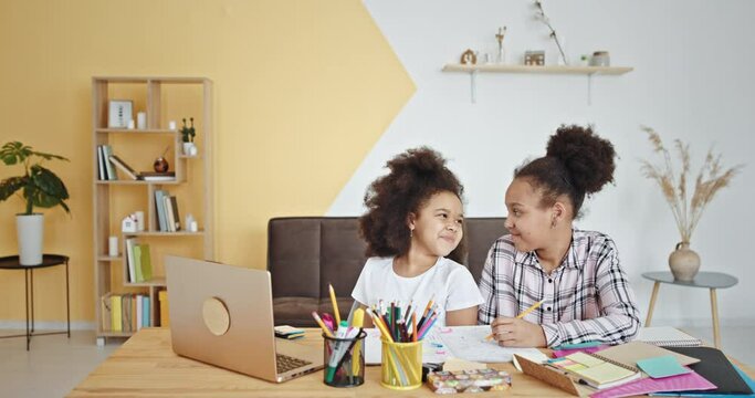 Black sisters doing homework together. Mixed race girls with curly hair smiling and doing homework assignment in copybook while sitting at table and studying at home together