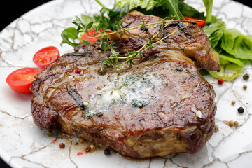 Grilled meat steak with butter, herbs and cherry tomatoes on a white plate, against a dark background
