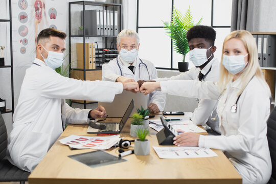 Fout multicultural medical experts in face masks and lab coats greeting with fists on meeting. Team of doctors sitting at table with various modern gadgets and looking at camera.