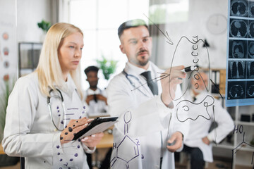 Obraz premium Team of qualified medical scientists using tablet and writing on glass flip chart during meeting at hospital room. Clinical studies and cooperation concept.