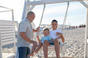 Happy father swings his sons on a swing by the sea. Big family on vacation on the coast in summer